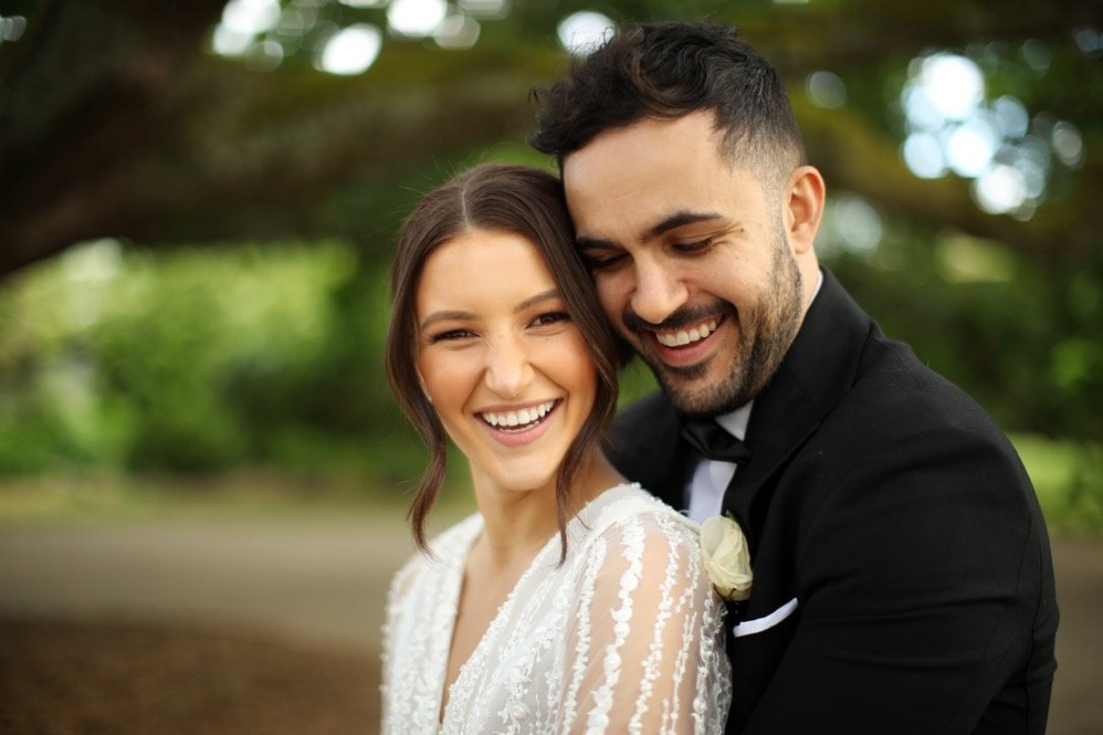 A smiling bride and groom partaking in a joyful moment outdoors, dressed in wedding attire, with a blurred natural background.