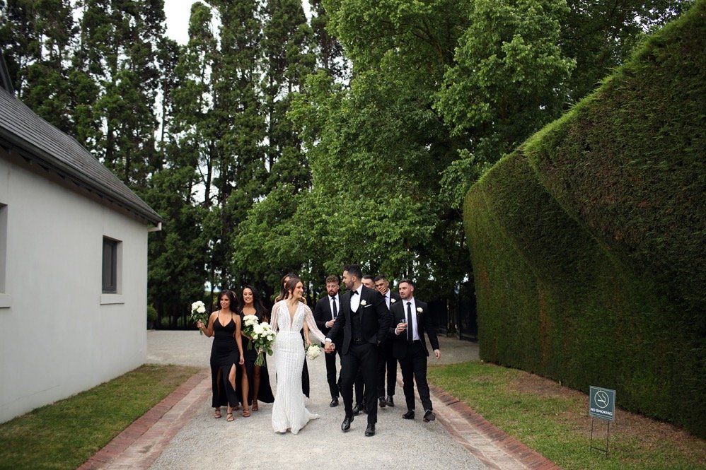 A wedding party walking outdoors on a gravel path, including a bride in a white dress, a groom in a black tuxedo, and several bridesmaids and groomsmen in formal attire, with trees and neatly trimmed hedges in the background.