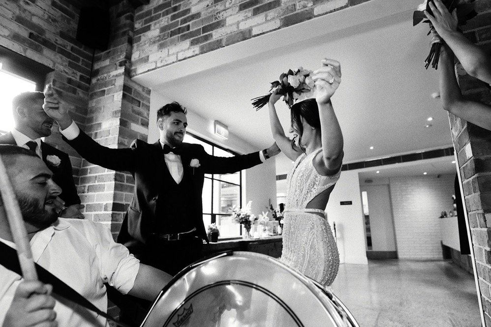A joyful bride holding a bouquet celebrates with her friends at a wedding reception, with a drummer in the foreground and friends in suits cheering around her.