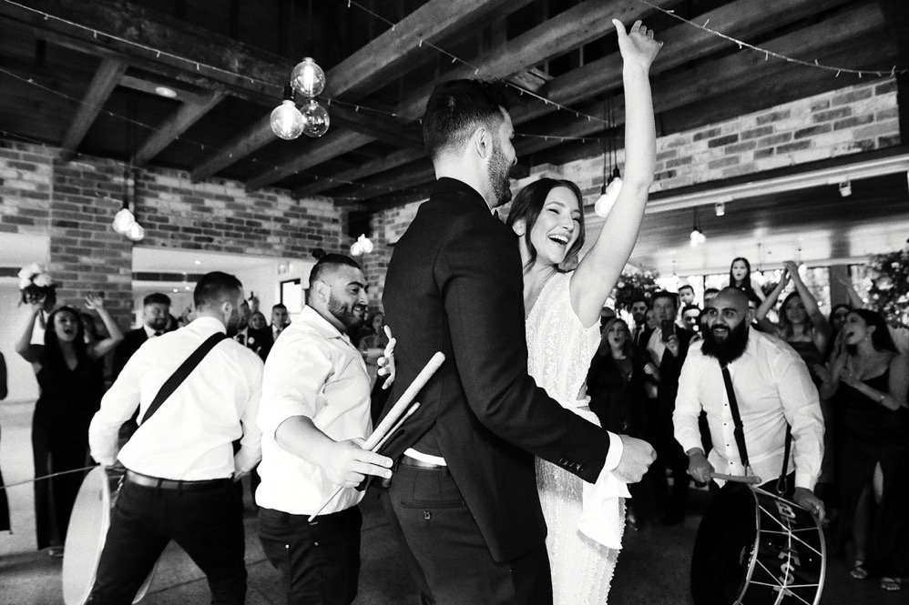 A wedding reception with a bride and groom dancing, surrounded by friends who are celebrating and playing musical instruments in a rustic, brick-walled venue.