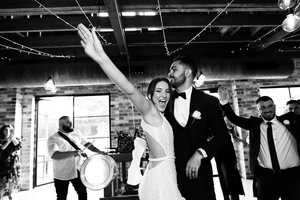 Black and white photo of a wedding reception with a bride and groom dancing together, smiling and celebrating. The bride is wearing a sleeveless wedding gown with cutouts, and the groom is in a tuxedo. Other guests are dancing around them, with some