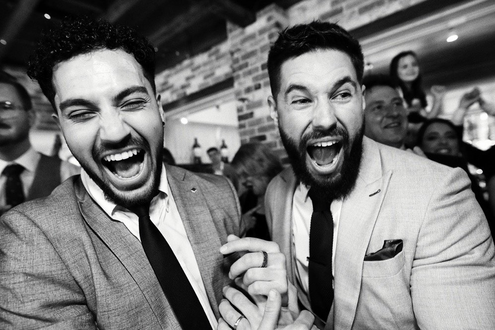 Two men in suits laughing and celebrating at a social gathering or party with a brick wall background.