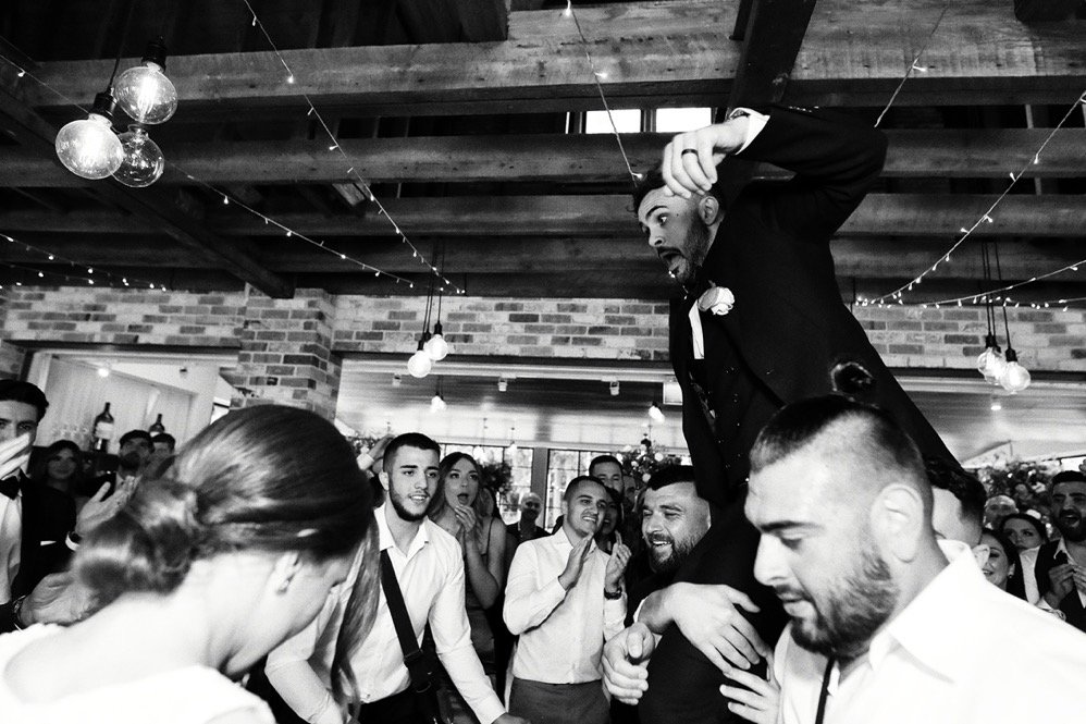 Man in tuxedo being lifted on shoulders during celebration, surrounded by dancing group in a rustic indoor venue with hanging lights and brick walls
