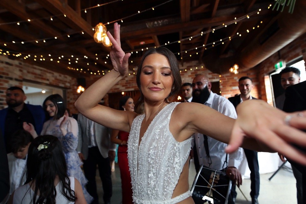 A woman dancing at a celebration or wedding with people in the background, decorated with string lights and brick walls.