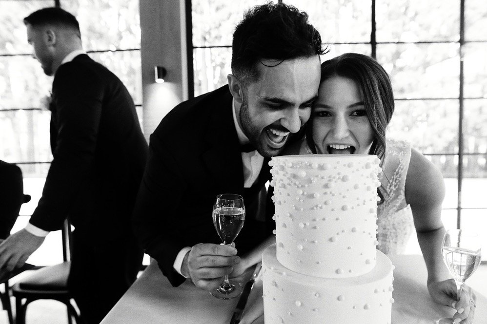 A couple celebrates at a wedding reception, cutting a two-tier white wedding cake with white decorations, each holding a glass of champagne, with a man in the background.