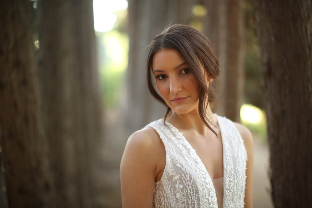 A woman with dark hair in a loose updo wearing a white lace sleeveless dress, standing outdoors between trees.