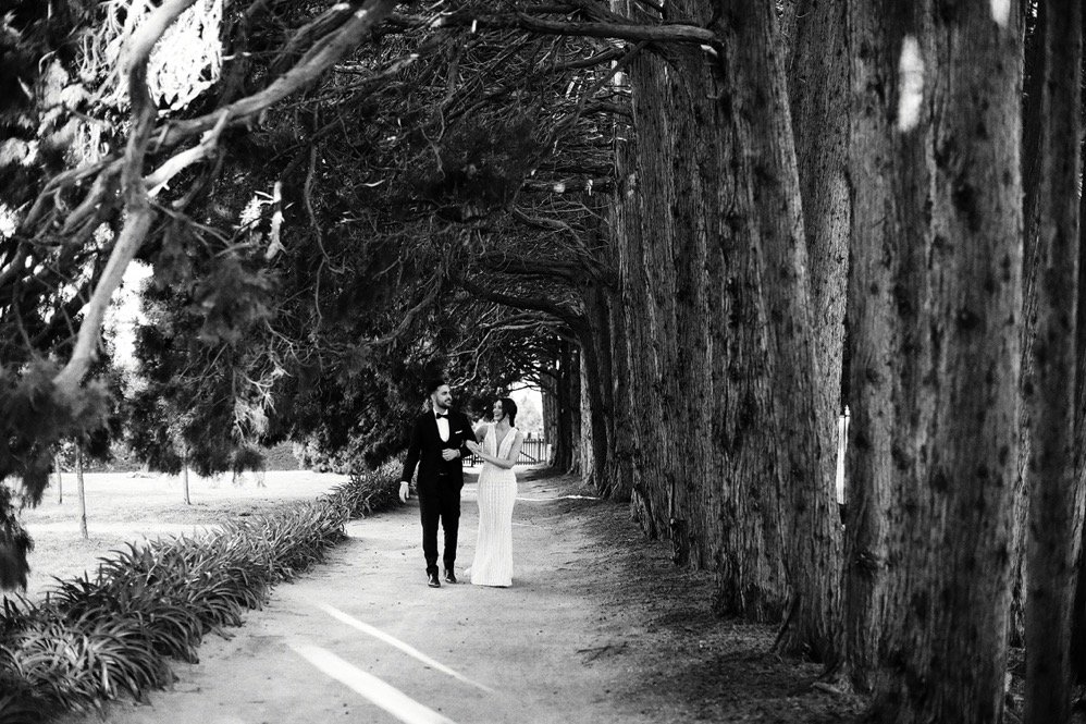 A couple dressed in formal attire, walking together along a tree-lined path in a park or garden.