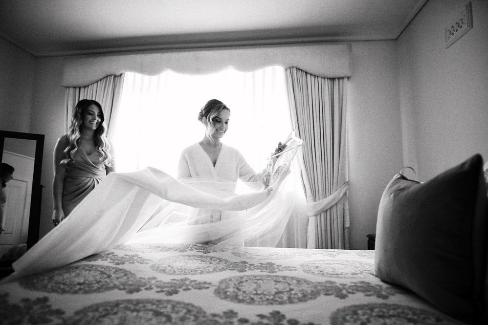 A woman in a white robe preparing a wedding dress on a bed while another woman looks on in a bright, elegant room with curtains and a large mirror.