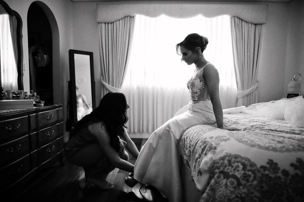 Woman in wedding dress sitting on bed, another woman helps her with her shoe in a bedroom with a dresser, mirror, and window with curtains.
