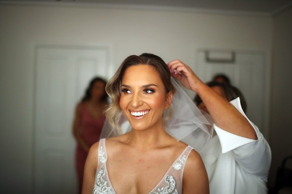 Bride smiling as a man adjusts her hair in a room, with two women blurred in the background.
