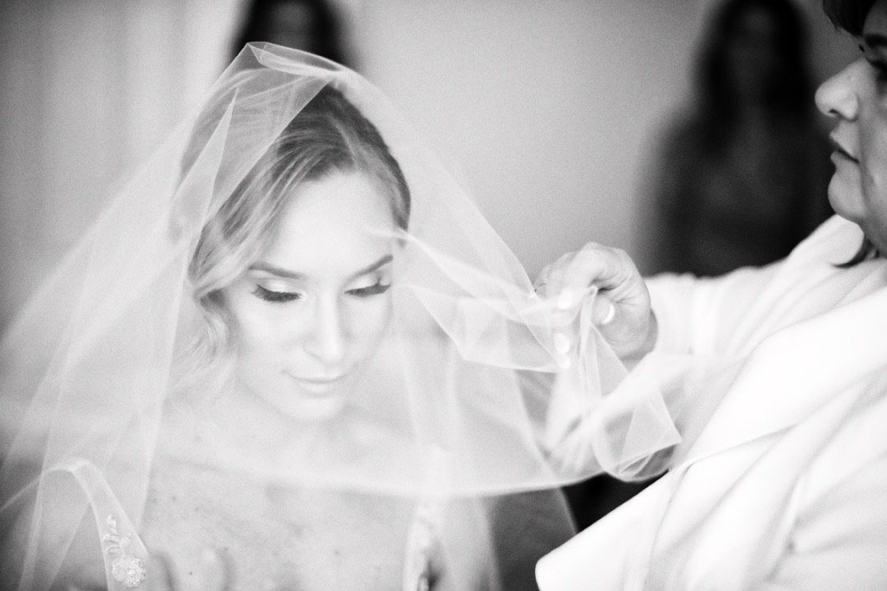 A bride with makeup and a veil, having it adjusted by a woman in white attire.