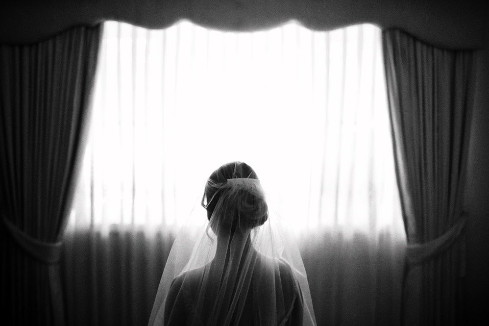 A woman in a wedding dress and veil stands facing a window with curtains, backlit in black and white.