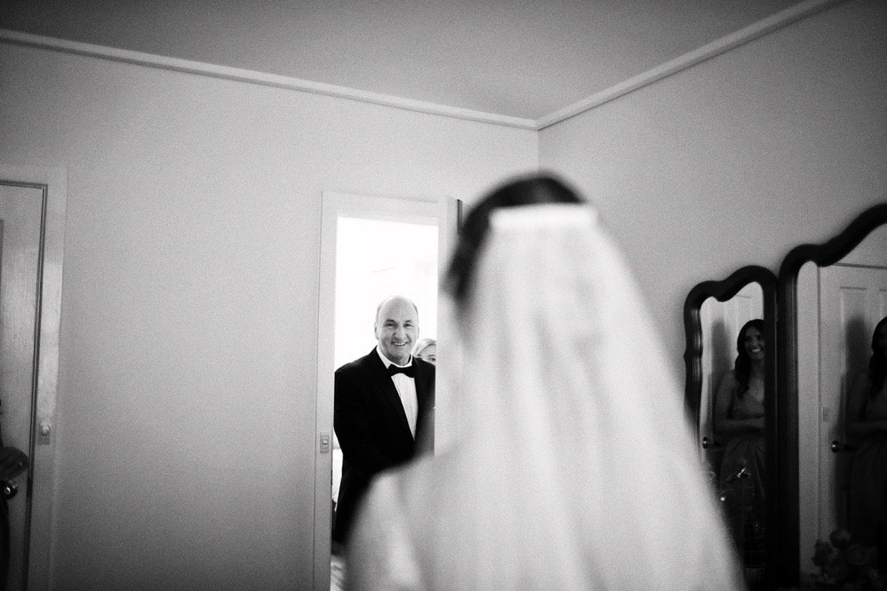 A bride in a wedding veil is in the foreground, while an older man in a tuxedo is smiling in a doorway in the background.
