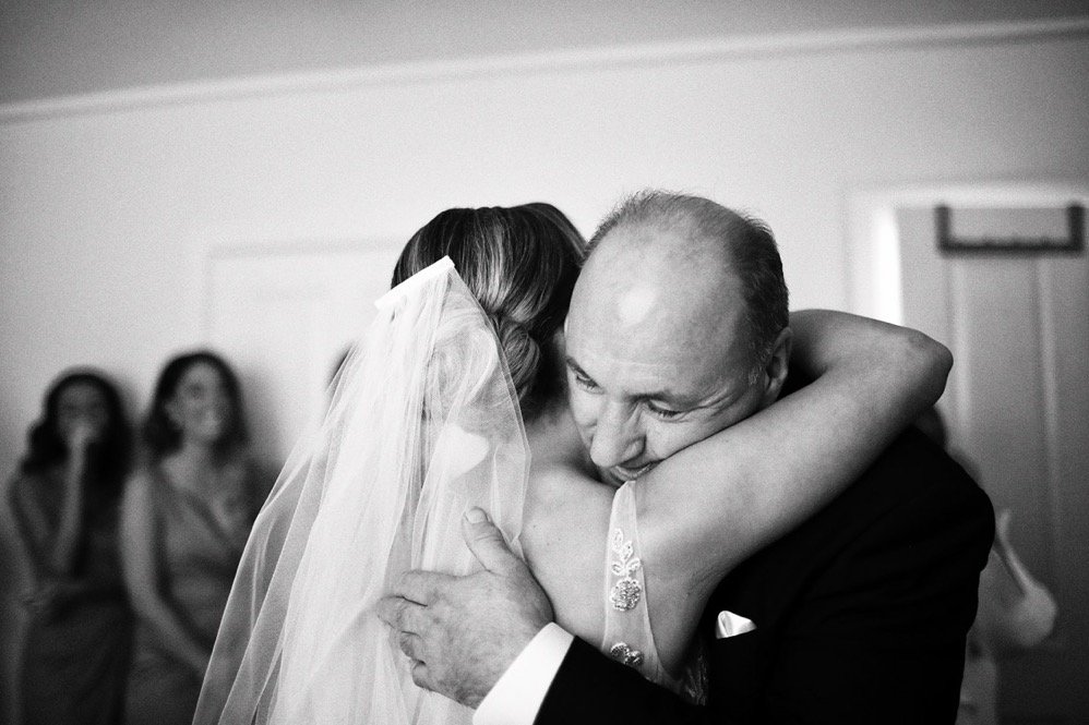 A bride and an older man, possibly her father, sharing a heartfelt hug during a wedding, with two women in the background watching.