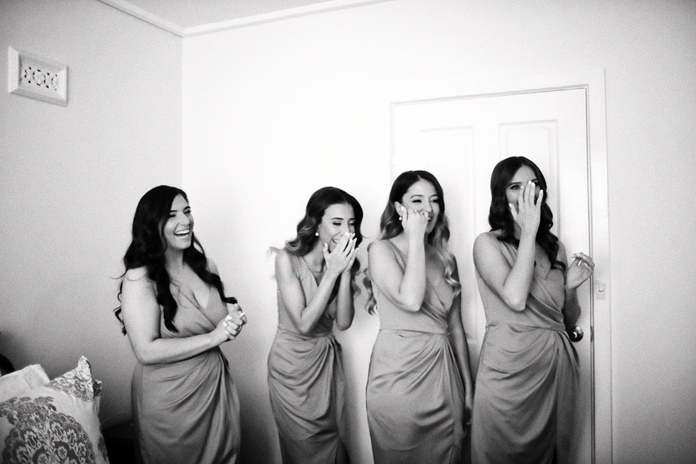 Four women in matching dresses laughing and covering their mouths in a room with plain walls and a closed door.