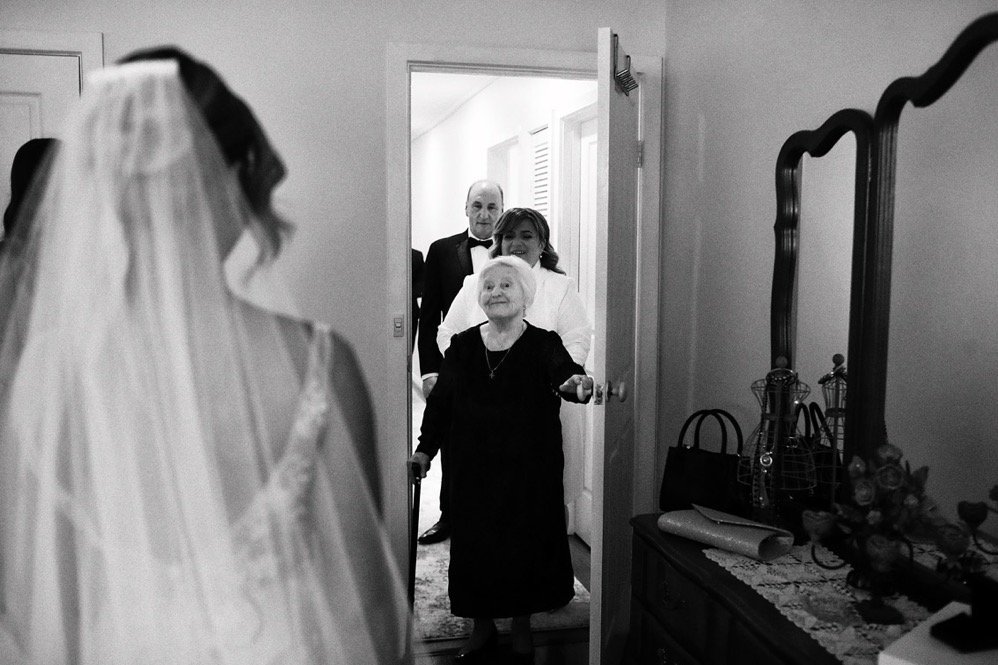 A bride in wedding attire looking at four guests, including an elderly woman with a walker, in a bedroom.