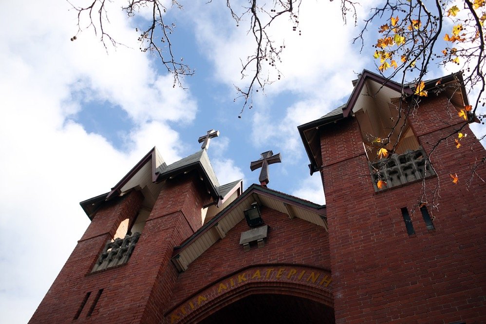 A red brick church with three crosses on its roof, viewed from below against a cloudy sky and tree branches.