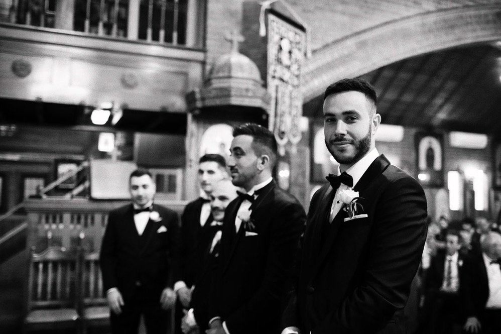 A group of men in tuxedos attending a wedding in a wood-paneled venue, with one man in the foreground looking at the camera.