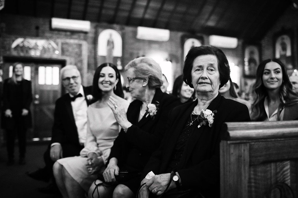 Black and white photo of people sitting inside a church during a wedding, with women wearing floral corsages, and one woman in front looking directly at the camera.