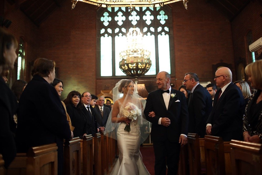 A bride walking down the aisle with her father at her wedding, inside a church with stained glass windows and a chandelier overhead.