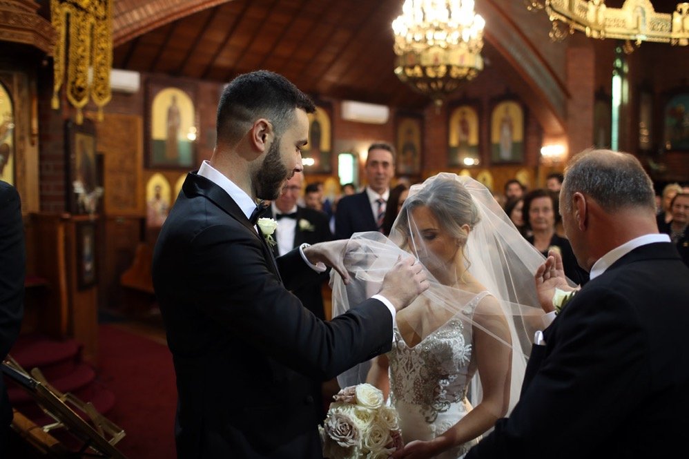 A groom placing a wedding ring on the bride's finger during a wedding ceremony inside a church, with guests observing in the background.