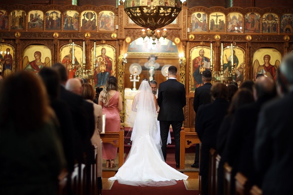 A wedding ceremony inside a church with a bride and groom standing before the officiant, surrounded by seated guests and religious icons on the wall.
