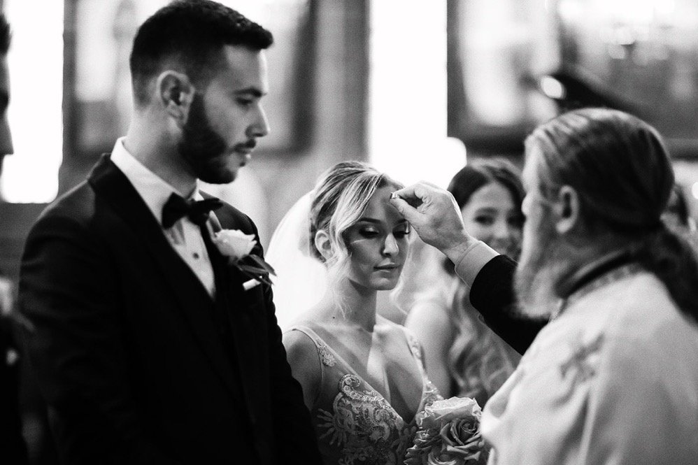 A bride and groom are having a wedding ceremony in a church, with a priest touching up the bride's forehead.