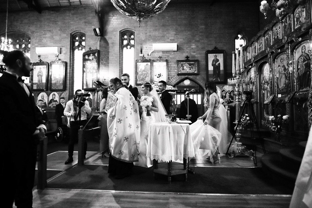 A wedding ceremony inside a church with a priest conducting the service, the bride in a white dress holding a bouquet, guests dressed in formal attire, religious icons on the walls, and a photographer capturing the moment.