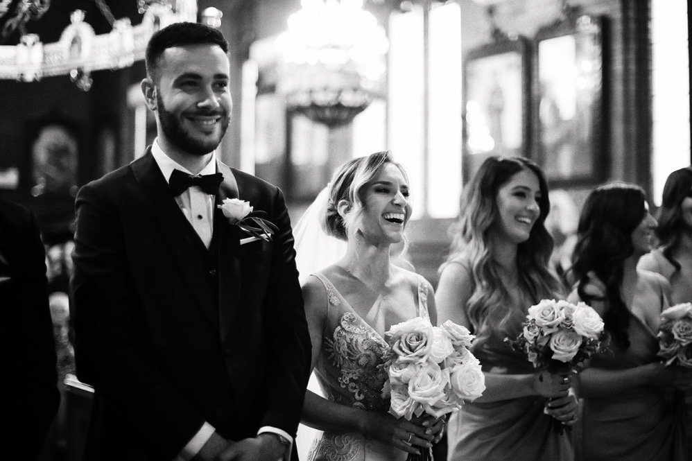 People in wedding attire holding bouquets, smiling, at a wedding ceremony in a church.
