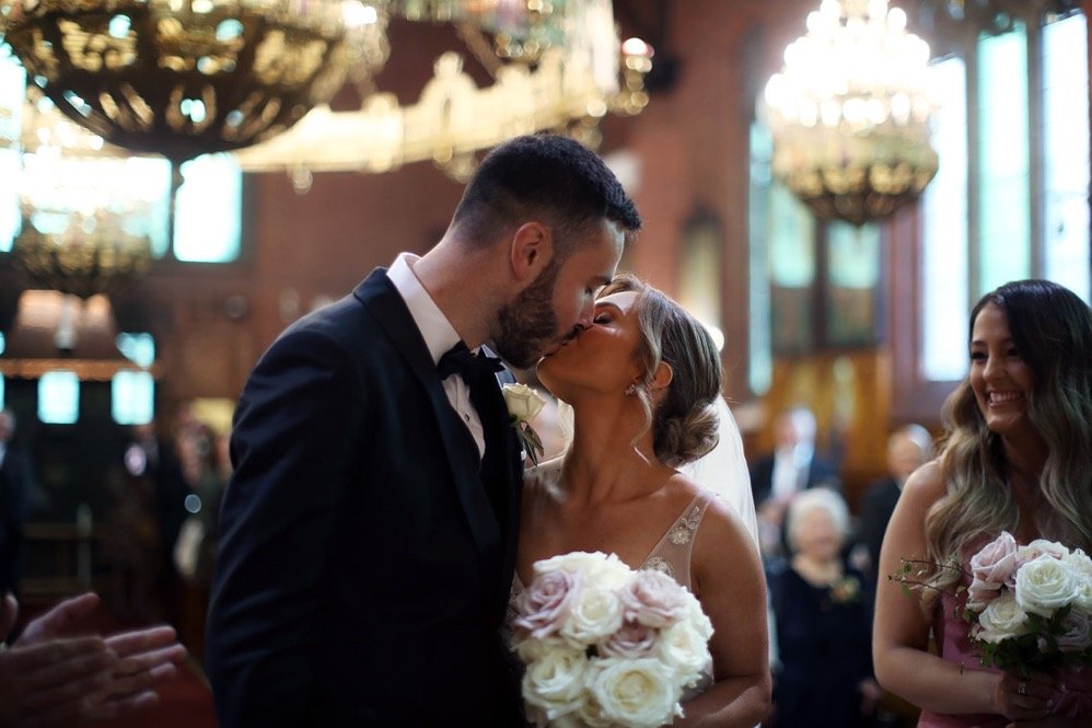 A groom and bride sharing a kiss during their wedding ceremony inside a church, with bridesmaids and guests present.