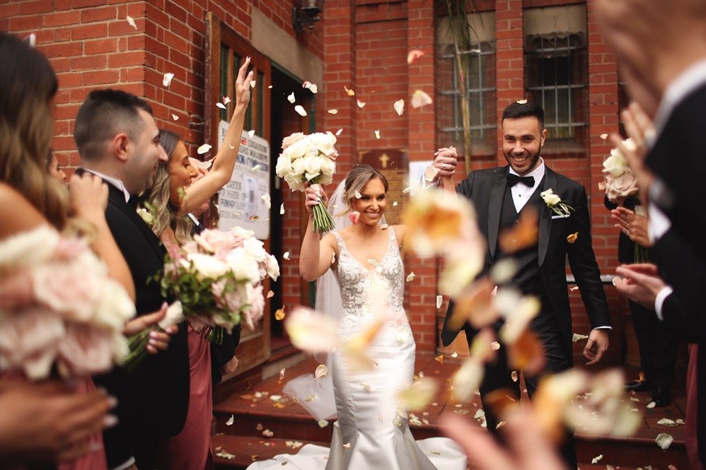 A bride and groom are celebrating their wedding surrounded by friends, throwing flower petals in the air.