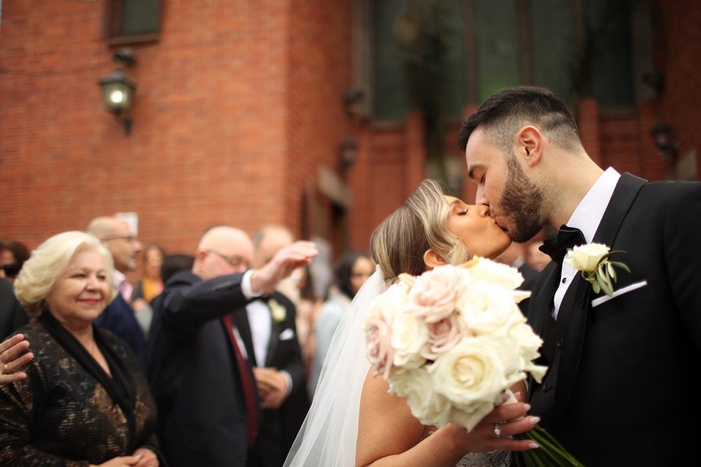 A newlywed couple sharing a kiss outside a brick building during their wedding, with guests watching in the background.