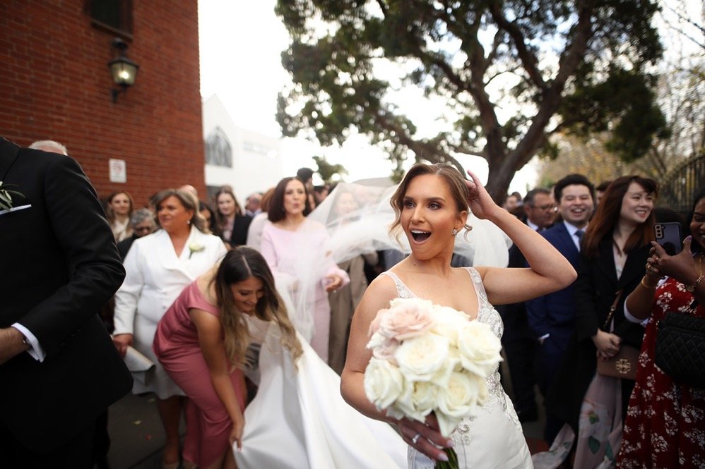 Bride holding a bouquet of white roses, smiling, in a wedding celebration with guests around her outdoors.