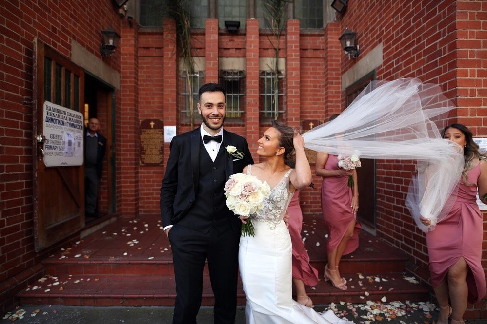 Happy bride and groom standing outside a red brick church, with bridesmaids in pink dresses throwing flower petals