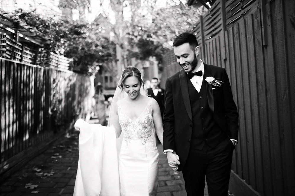 Black and white photo of a bride and groom walking hand in hand outdoors, smiling, with the bride in a lace wedding dress and the groom in a tuxedo.