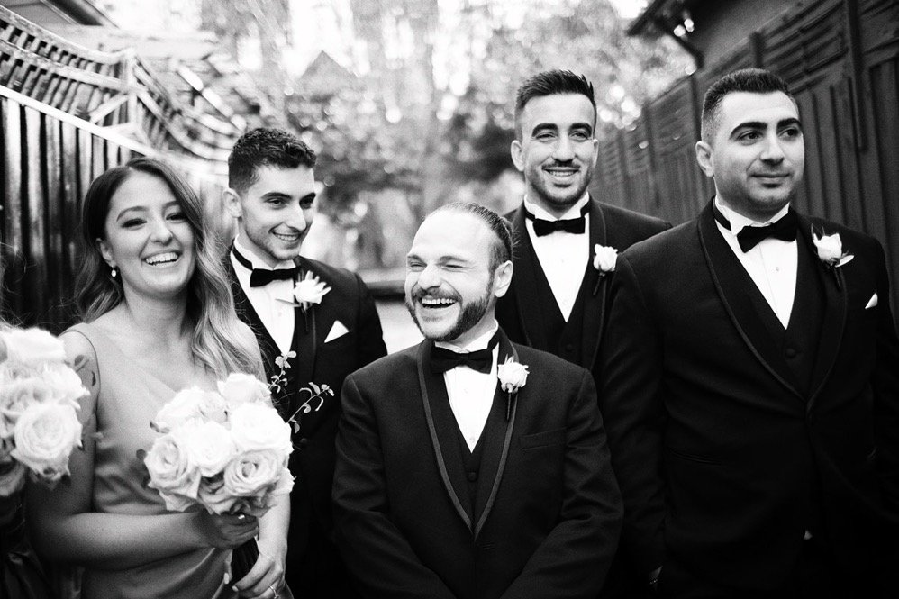 Black and white photo of five people dressed in formal attire, likely at a wedding. The woman on the left holds a bouquet of roses. The man in the center has a beard and is smiling broadly. The other three men and woman are also smiling, dressed in t