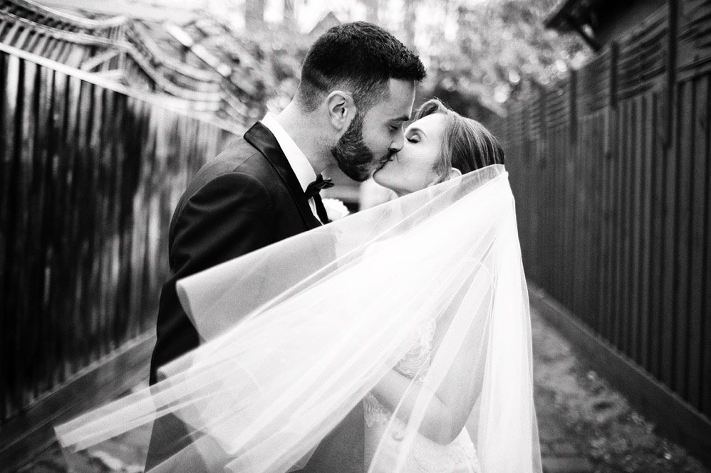 Black and white photo of a bride and groom sharing a kiss, with the bride's veil flowing around them, in an outdoor setting.