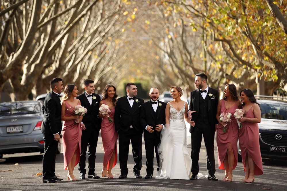 Wedding group photo of eleven people standing on a street with trees and parked cars, including a bride and groom and seven bridesmaids and groomsmen, during autumn.