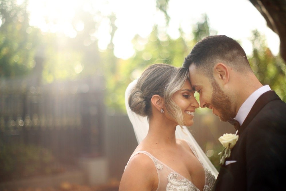 A bride and groom with foreheads touching, smiling, outdoors during daytime, with sunlight and blurred trees in the background.