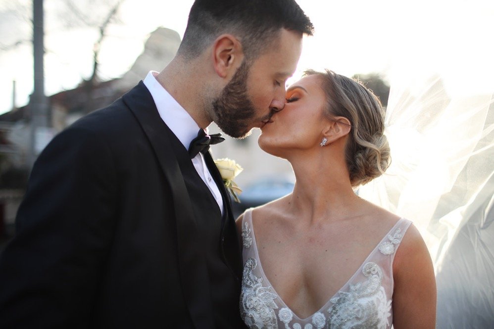 A bride and groom sharing a kiss outdoors on their wedding day, with the bride wearing a white gown and veil, and the groom in a black tuxedo.
