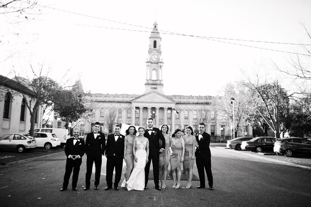 A group of nine people, dressed in formal attire, standing outside in front of a historic building with a clock tower, trees, and parked cars on the street.