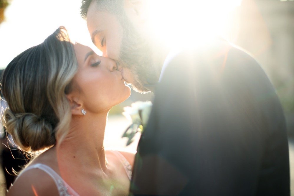 A couple sharing a kiss outdoors with the sun shining behind them, creating a bright glow around their faces.