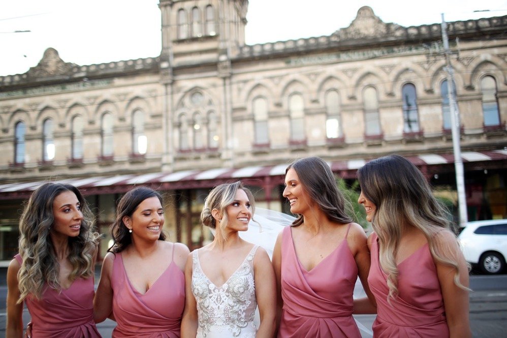 A bride and four bridesmaids stand together outside a historic building, smiling and looking at each other during a wedding celebration.