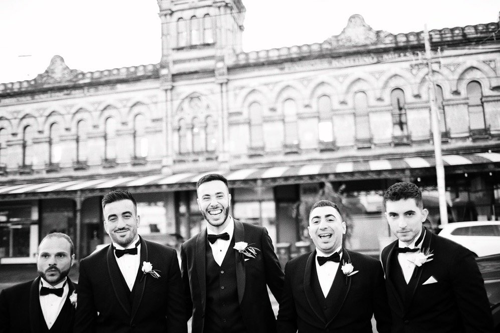 A group of five men dressed in tuxedos with boutonnières, standing outside a historic building, smiling and posing for a photo.