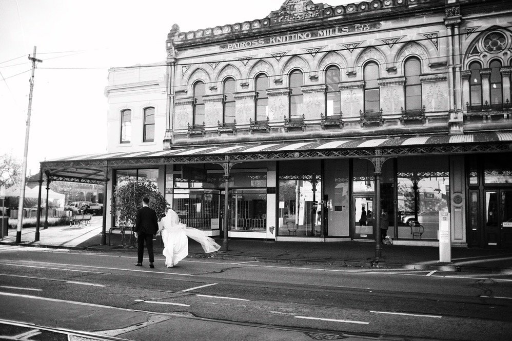 A bride and groom walking in front of a historic building with the sign "Patross Knitting Mills" on the roof, in black and white.