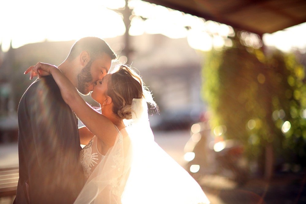 A couple, likely newlyweds, embracing outdoors during sunset, with the bride in a wedding dress and veil, and the groom in a suit, sharing an intimate moment.