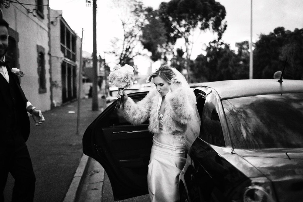 A bride exiting a car, holding a bouquet, wearing a fur coat over her wedding dress, with one hand adjusting her veil, while a man in a suit stands nearby on the sidewalk in a black and white photo.