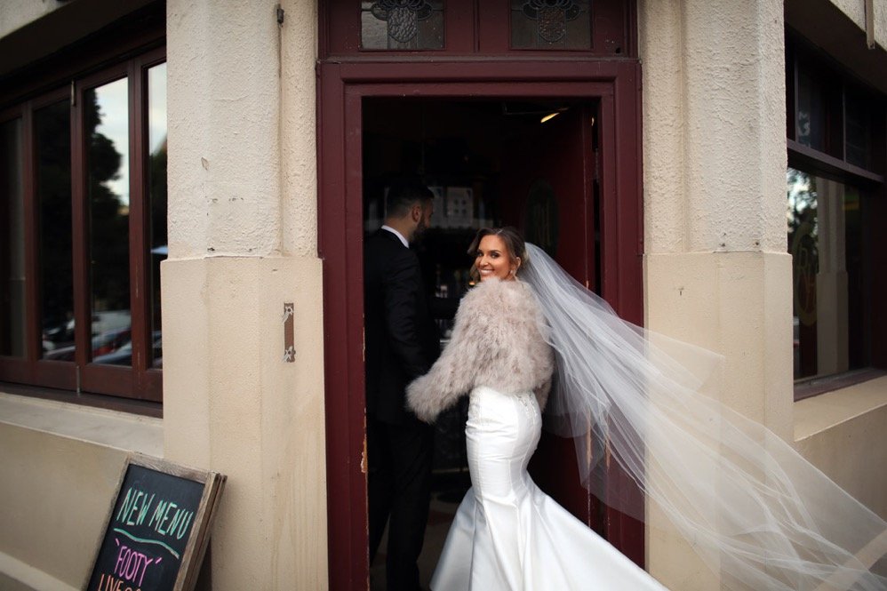 A bride in a white gown and veil smiling while looking back at the camera outside a doorway, with a groom in a black suit inside the building.