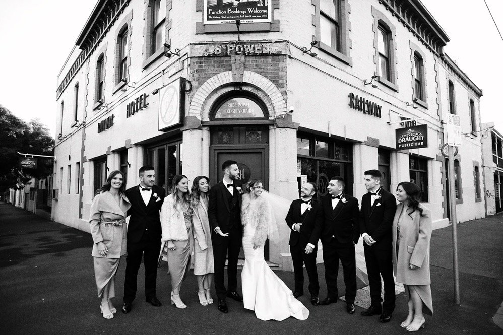A wedding party with the bride in a white gown and veil, surrounded by groomsmen and bridesmaids in formal attire, standing outside a corner building with signs for a hotel, pub, and public bar.