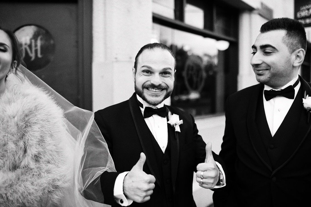 Two men in tuxedos smiling and giving thumbs up at a wedding, with one man wearing a boutonniere.